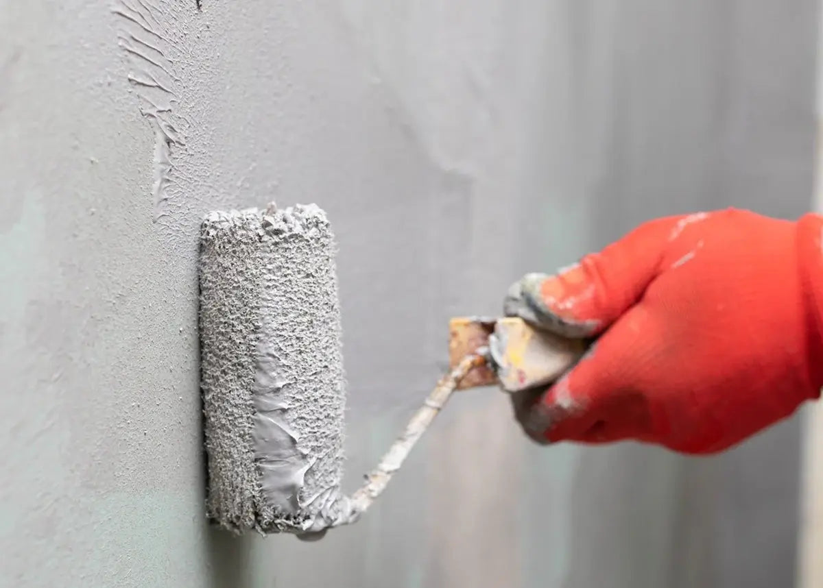 Hand with a roller applying gray waterproofing to a wall