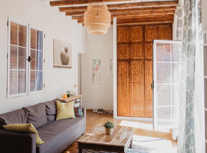 Interior living room scene with a gray couch, wooden door, and a painting on the wall. Natural lighting and a potted plant on the floor add to the scene.