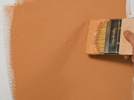 A close-up of a paintbrush spreading ochre-colored cork paint onto a wall.