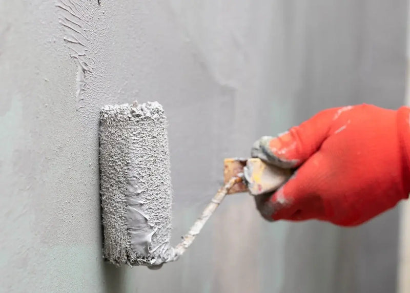 Hand with a roller applying gray waterproofing to a wall