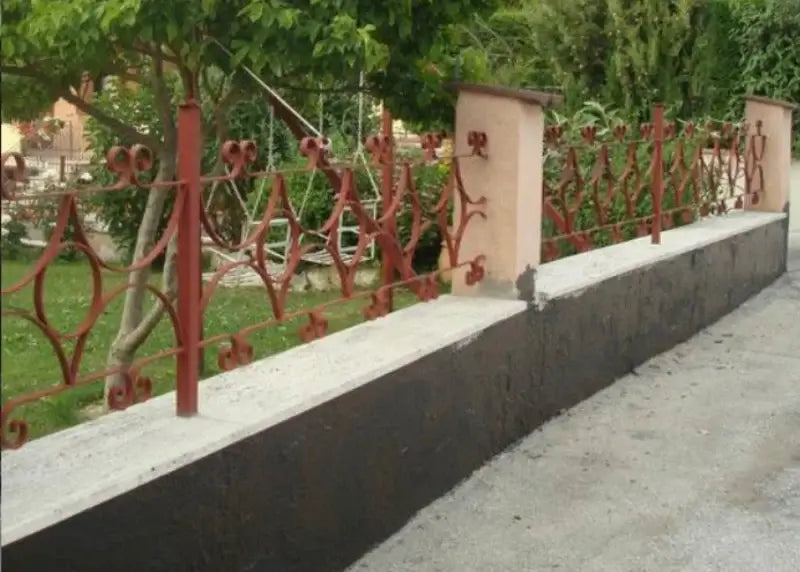 Red metal fence with decorative elements on a concrete wall, greenery in the background